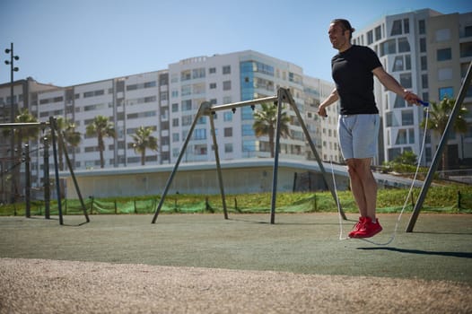 Active young athletic man warming up his body, exercising with a jumping rope on the outdoor urban sportsground