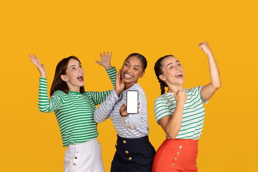 Emotional young ladies girlfriends showing smartphone with white blank screen mockup copy space gesturing and exclaiming, celebrating beginning of season sale, yellow background