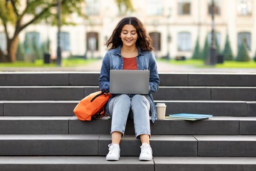 Smiling indian student woman sitting on stairs working on laptop, preparing for exams outdoors, having rest in university campus. Technology, education and remote working concept, copy space