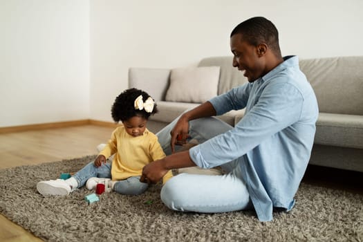 Playtime. Cheerful loving african american father playing colorful toys with his little daughter, girl moving cubes, spending free time together at home interior, copy space