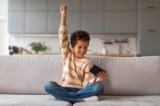Joyful black boy holding smartphone and raising his arm in victory