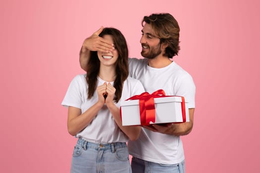 Delighted woman with eyes covered by a man's hand, receiving a surprise gift box with a red ribbon