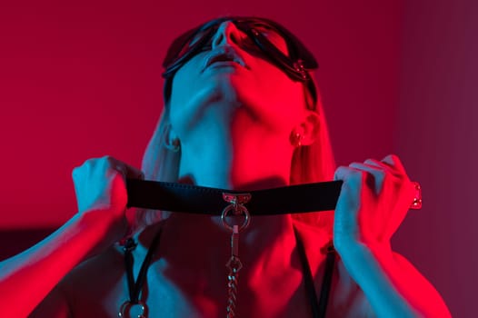 Blonde woman in leather mask putting on collar in bedroom with red blue neon light.