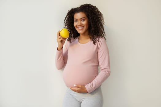 Beautiful young black pregnant woman with apple in hand standing against white wall background, happy african american expectant mother tenderly touching belly and smiling at camera, copy space