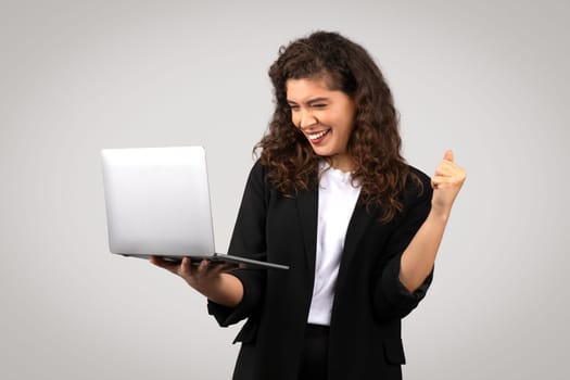 Cheerful businesswoman with curly hair looking at laptop and raising her fist in victorious gesture, symbolizing success and achievement, grey background