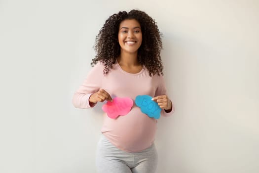 Gender Reveal Party. Happy black pregnant woman holding pink and blue cutout cards with 'GIRL' and 'BOY' text, african american expectant mother expressing excitement for upcoming motherhood