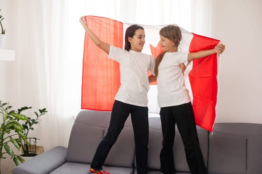 Two sisters are sitting on a couch at home with canadian flags on hands. Canada children girls with flag . High quality photo