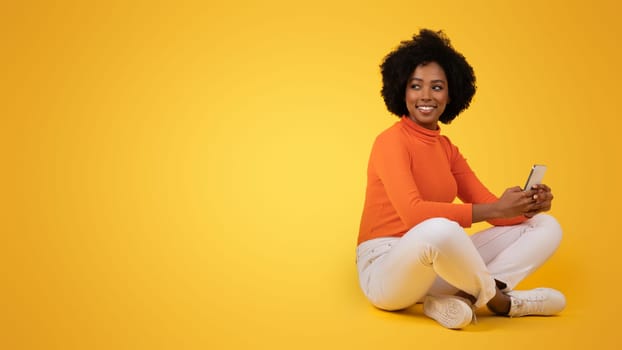 Content African American woman with curly hair sitting cross-legged, holding a smartphone