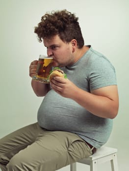 Man, drink and beer with burger in studio for unhealthy diet, fast food and nutrition on chair. Plus size, hungry and male person with alcohol on stool for dinner, meal and beverage in glass.