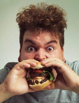 Takeaway, burger and food for man in studio with meal for big eater, plus size or greed. Young person with meat in mouth for hunger, silly and bite indoor for delicious, taste and sauce for portrait.