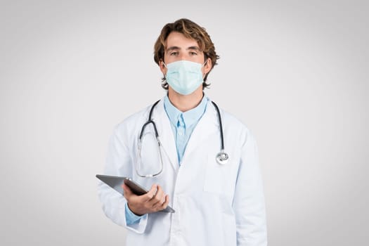 Focused male doctor wearing surgical mask and stethoscope, using digital tablet, signifying healthcare and technology fusion, grey background