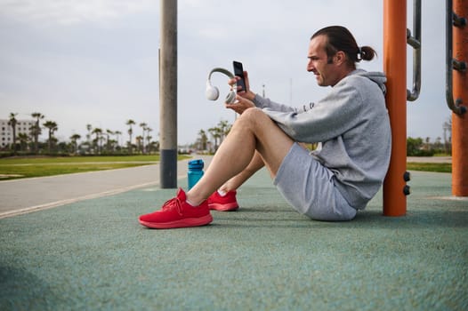 Athletic man checking mobile app on his smartphone while resting after workout the urban sports ground. Side view