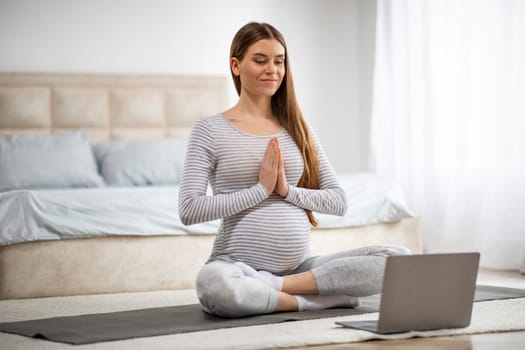 Calm Young Pregnant Woman Having Online Yoga Lesson With Laptop At Home, Beautiful Expectant Mother Meditating In Front Of Computer, Sitting In Lotus Position On Mat And Making Namaste Gesture