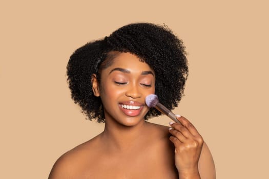 Joyful black woman applying blush, makeup routine joy