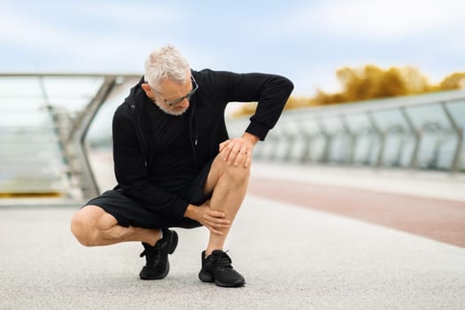 Elderly sportsman exercising in public park, touching his injured leg