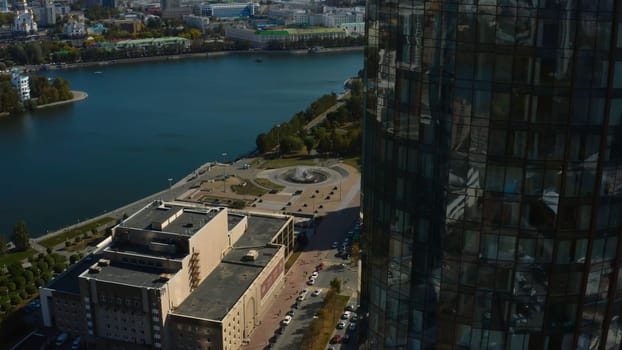 Top view of glass facade of high-rise building with landscape of city and river. Stock footage. Beautiful landscape of modern city with glass buildings. Glass skyscrapers with green landscapes of summer city with river