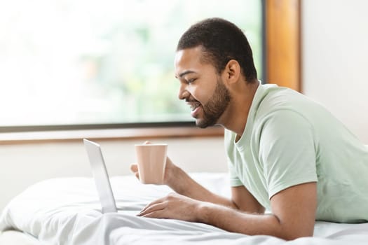 Man lying in bed using laptop and holding cup