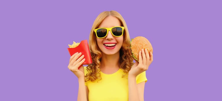 Portrait of happy cheerful young woman eating burger fast food and french fries, fried potatoes isolated on purple studio background