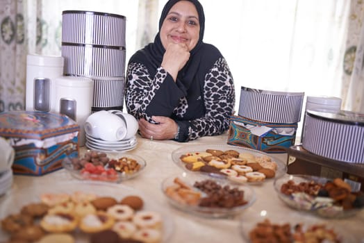 Mature woman, Islamic and food in house with cheerful, smile and dining table for Iftar. Muslim person, cookie dessert and joy with Eid Mubarak, religion and celebration for breaking fast at home