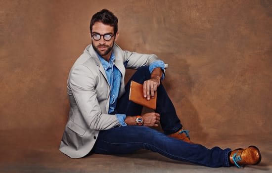 Fashion, book and man with glasses in studio with trendy, stylish and classy suit on floor. Reading, story and handsome male person with elegant style for outfit and novel by brown background.