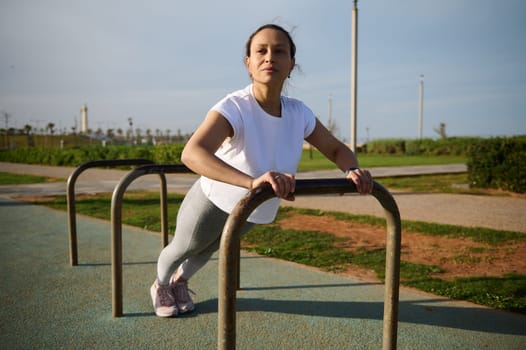 Strong athletic young woman working out, training arms while doing push ups on crossbar in the outdoor sports ground