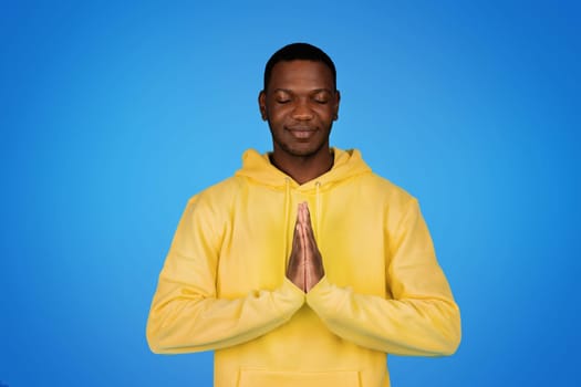Smiling millennial african american guy prayer in hoodie with eyes closed making pray sign