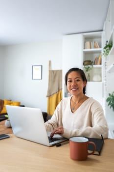 Happy Asian female entrepreneur working using laptop looking at camera. Smiling Chinese woman working at home office.