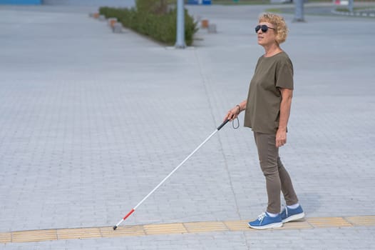 An elderly blind woman walks with a cane along a tactile tile.
