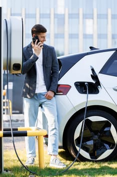 Handsome businessman calling while charging his electric car on the street. Portrait of joyful 25-aged man which has mobile conversation while electric car charging battery on charging station