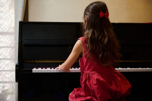Back view of a cute child girl, a pianist student, future musician pianist touching the white keys with his fingers to create the rhythm of the melody, enjoying playing grand piano during music lesson