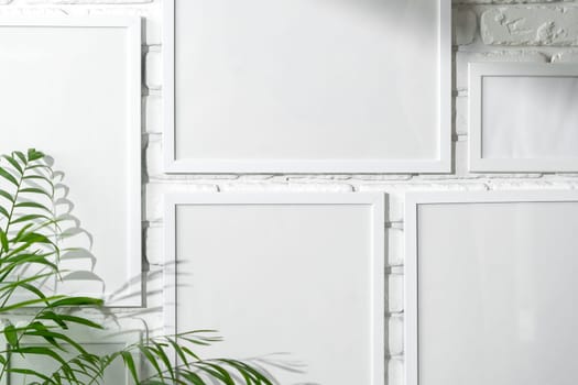 Several empty photo frames of different sizes and colors are displayed in an asymmetrical arrangement against a textured white brick wall, potentially signifying a modern and minimalist interior design approach. A small portion of a green plant is visible, injecting a touch of nature into the scene.