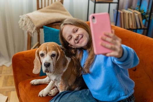 POV of smiling girl child kid with cute beagle dog pet taking selfie on smartphone at home room
