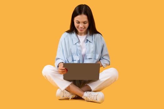 Young lady with laptop sitting cross-legged on yellow background