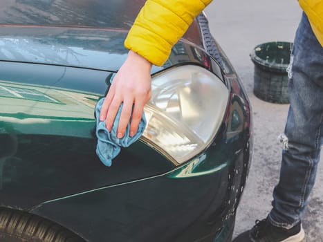 A young caucasian guy in a yellow jacket holds blue rags in his hands and wipes the headlight of his car.