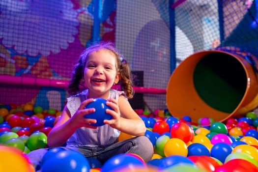 A young girl with a beaming smile sits amongst a sea of vibrant multicolored balls in an indoor play area. Her casual attire suggests a day of fun and activity as she reaches out, enjoying the playful environment around her. The background features soft play structures and a tunnel slide, highlighting the energetic atmosphere of the childrens playground.
