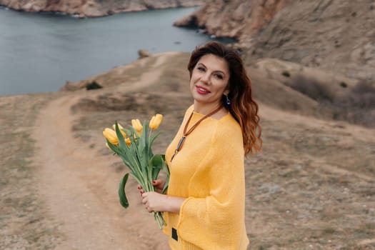 Portrait of a happy woman with long hair against a background of mountains and sea. Holding a bouquet of yellow tulips in her hands, wearing a yellow sweater.
