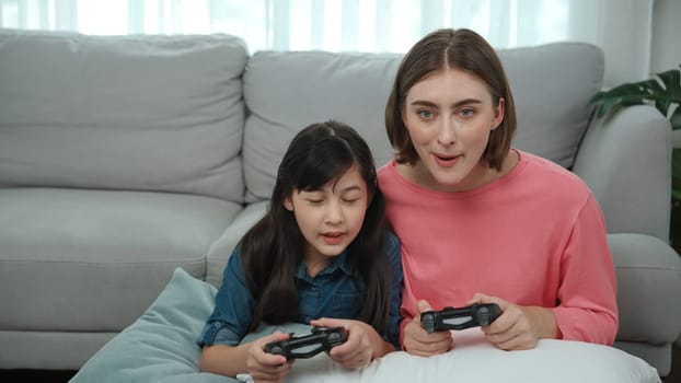 Caucasian mom and daughter sitting and playing game at living room. Happy mother spend time together with smart asian girl while holding joystick ans enjoy playing games. Lifestyle concept. Pedagogy.