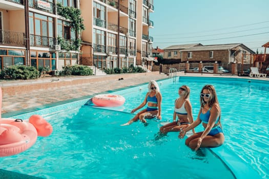 Three women are sitting in a pool with pink and blue floaties. Scene is lighthearted and fun.
