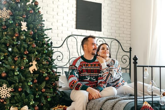 Young woman and man sitting on bed in sweaters and hugging near Christmas tree