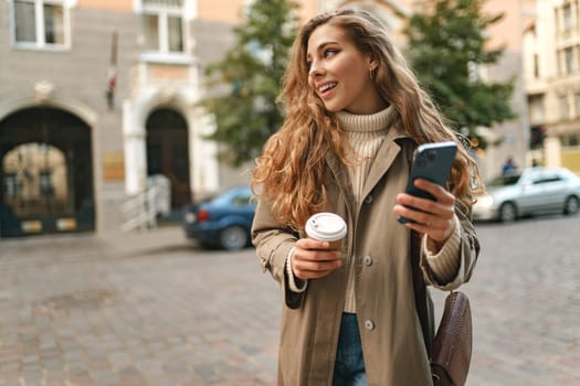 Young woman wearing autumn coat walking with smartphone and coffee cup in a city street