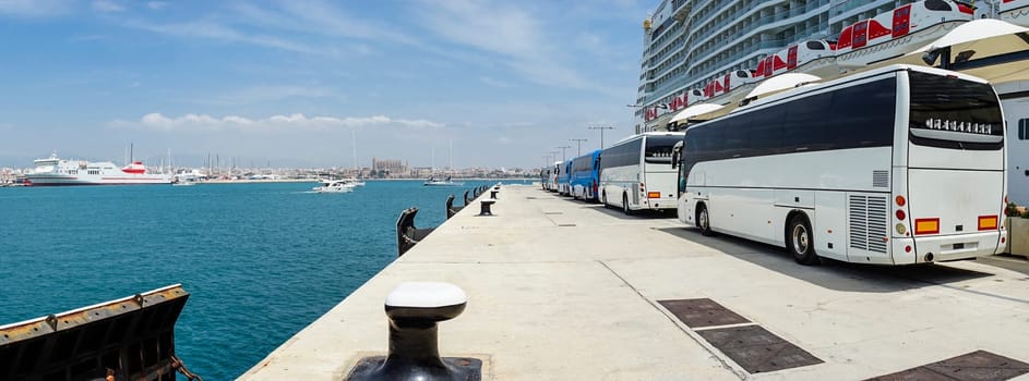 Tour buses await passengers near a cruise ship at a busy port, with a scenic cityscape and clear blue sea