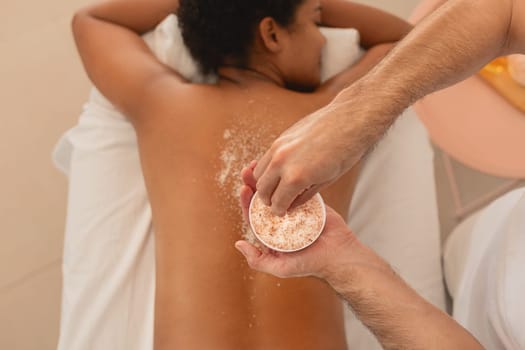 A practitioner is seen applying a scrub to the back of a client African American woman lying on a spa table, indicating a spa treatment scenario