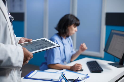 Close-up shot of medical professionals utilizing modern technology for discussing healthcare in a clinic. Selective focus shows a person holding a tablet and a female nurse using a computer.