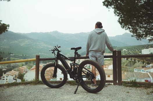 Rear view of a young active man cyclist standing near his electric battery powered bike, admiring beautiful mountains, enjoying riding in the nature