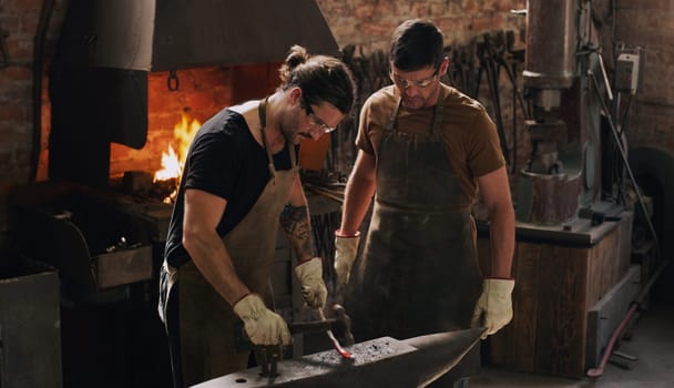 Blacksmith, colleagues and hammer with metal, flame and equipment for professional craft. Protection, teamwork and gloves in workshop with anvil for metalwork, forge and iron product creation factory