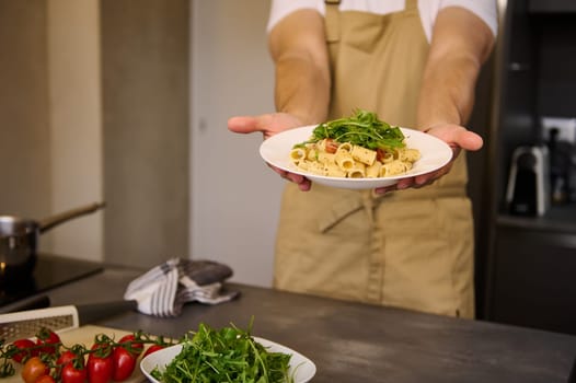 White plate with Italian pasta, garnished with tomato sauce and fresh arugula leaves in the hands of male chef holding out at camera the dish, standing at kitchen table with ingredients. Food concept
