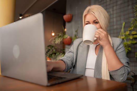 A woman is sitting at a table with a laptop and a white coffee cup. She is drinking coffee while working on her laptop.