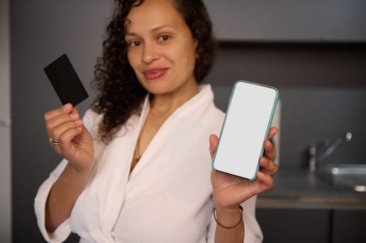 Young woman holds and shows at camera blank black credit card and smartphone with white empty mockup digital screen, makes online payments, transfers money via internet mobile banking or application