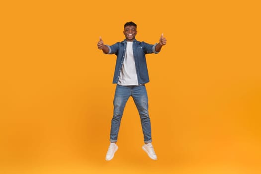 A young black man with a joyful expression jumps with thumbs up, representing atmosphere of happiness, isolated on an orange backdrop