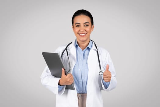 Cheerful latin woman doctor approving service, showing thumb up and smiling at camera, posing on grey background